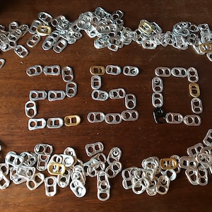 May include: A display of silver and gold-coloured can tabs forming the number "250" on a wooden surface. The tabs are scattered around the numbers, creating a decorative arrangement. One black tab is used in the number.