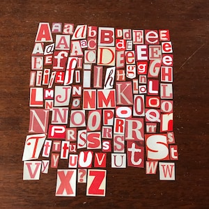 Puede incluir: Una colecci&oacute;n de letras del alfabeto de papel rojo y blanco, recortadas de revistas o peri&oacute;dicos, dispuestas en orden alfab&eacute;tico sobre una superficie de madera marr&oacute;n.