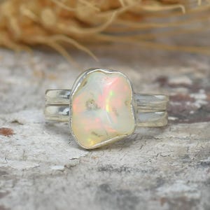 May include: A silver ring featuring a large, irregular-shaped opal stone with iridescent colors of pink, white, and yellow. The ring has a double band design. The background includes a textured surface and wheat stalks.