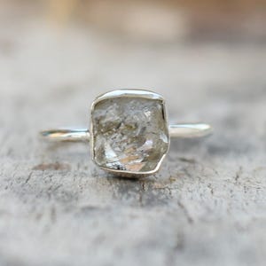 Könnte beinhalten: Ein Silberring mit einem quadratischen, roh geschnittenen, hellfarbigen Edelstein. Der Ring hat ein dünnes, poliertes Band, und der Stein hat eine leicht unebene Oberfläche. Die Fassung ist eine einfache Lünette. Der Ring wird auf einer strukturierten, hellfarbigen Oberfläche gezeigt.