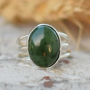 May include: A silver ring with a large, oval, dark green stone. The ring has a triple-band design. The stone is polished and reflects light. The background is blurred, with a neutral color palette.