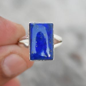 May include: A silver ring featuring a rectangular, deep blue lapis lazuli stone. The stone is set in a simple, elegant design. The ring is held up against a blurred background.