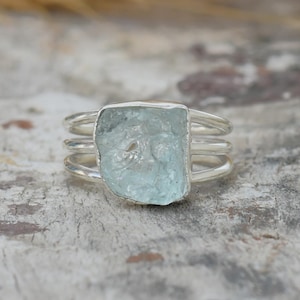 May include: A silver ring featuring a raw, light blue aquamarine gemstone. The ring has a triple-band design, with the stone set in the center. The background is a rustic, textured surface with hints of brown and gray.