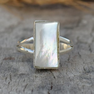 May include: A silver ring with a rectangular, iridescent white mother-of-pearl stone. The ring has a split shank design, with two bands merging to support the gemstone. The ring is set on a wooden surface.