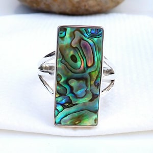 May include: A rectangular abalone shell ring with a silver band. The abalone shell has iridescent green, blue, and purple hues. The ring's band features a split design, adding a unique touch to the jewelry.