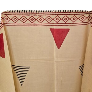 May include: Cream-colored textile with a geometric pattern. The design features red triangles and a border with diamond shapes. Black lines form triangles. The fabric is folded, showing the pattern's repeat.
