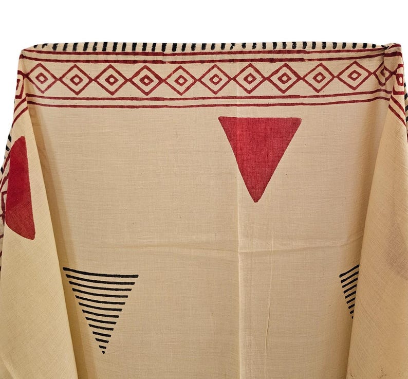 May include: Cream-colored textile with a geometric pattern. The design features red triangles and a border with diamond shapes. Black lines form triangles. The fabric is folded, showing the pattern's repeat.