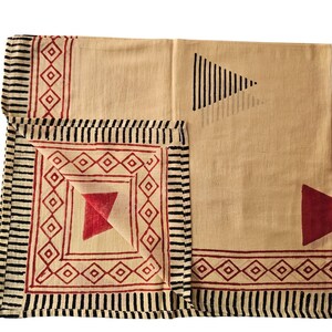 May include: Beige and red geometric patterned textile. The textile features a repeating diamond and triangle design in red, with black striped borders. The fabric is folded, showcasing the pattern variations. The overall design is symmetrical and visually appealing.
