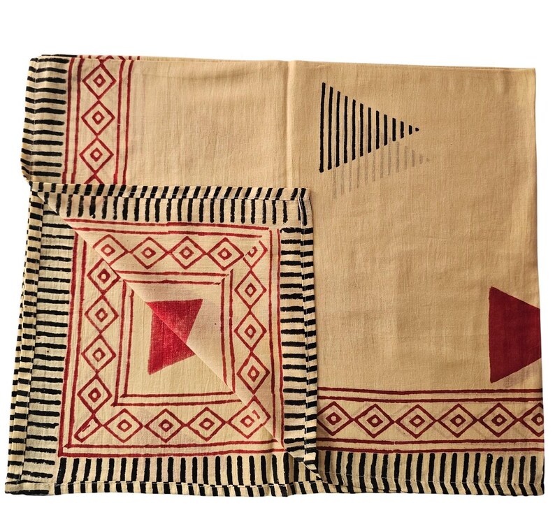 May include: Beige and red geometric patterned textile. The textile features a repeating diamond and triangle design in red, with black striped borders. The fabric is folded, showcasing the pattern variations. The overall design is symmetrical and visually appealing.