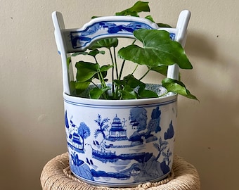 Puede incluir: Una maceta de porcelana azul y blanca con un diseño de paisaje chino, que presenta una planta con hojas verdes, sentada en un taburete de bambú tejido.
