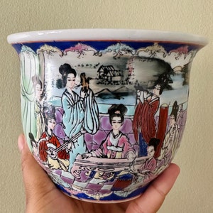 May include: A white ceramic planter with a blue rim and a colourful scene of women playing musical instruments. The scene is painted in a traditional Chinese style.