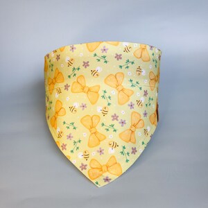 May include: Yellow bandana featuring a pattern of bees and bows. The bandana is made from a soft, lightweight fabric and is ideal for adding a touch of whimsy to any outfit.
