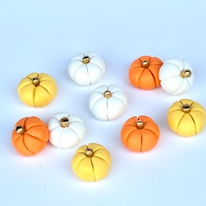 May include: A collection of small, decorative pumpkins in various colors, including white, orange, and yellow. Each pumpkin has a gold-colored stem. The pumpkins are arranged on a white surface, perfect for fall-themed crafts or decor.