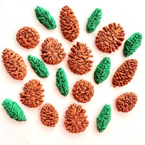 May include: A collection of brown pinecone-shaped items and green leaf-shaped items arranged on a white surface. The pinecones feature detailed, textured scales, while the leaves have a ribbed, pointed design. These decorative pieces are scattered.
