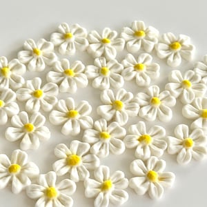 Könnte beinhalten: Eine Ansammlung kleiner, weißer Gänseblümchen-Blütenverzierungen mit gelben Zentren. Die Blüten sind auf einer weißen Oberfläche angeordnet und bilden ein Muster. Diese dekorativen Blumen eignen sich für Bastelprojekte, Scrapbooking oder zum Hinzufügen einer floralen Note.