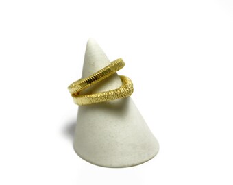 May include: A gold-toned ring with a textured, twisted design. The ring is displayed on a white cone-shaped stand.