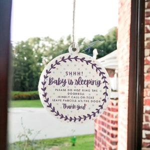 May include: A round, white sign with a purple border and a floral design. The sign reads "SHHH! Baby is sleeping. Please do not ring the doorbell. Kindly call or text. Leave parcels at the door. Thank you!"