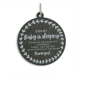 May include: A round wooden sign with a grey woodgrain finish and a white wreath design. The sign reads "SHHH! Baby is sleeping. Please do not ring the doorbell. Kindly call or text. Leave parcels at the door. Thank you!"