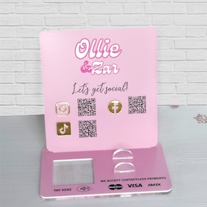 May include: Pink acrylic stand with the text "Ollie & Zar Let's get social!" and social media icons with QR codes. The stand also has a contactless payment area with the text "We accept contactless payments" and logos for Visa, Mastercard, and Amex.
