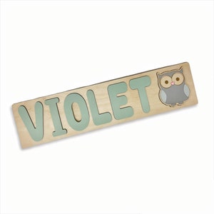 May include: Wooden name puzzle spelling "VIOLET" in light green letters. A light brown owl with gray and white accents is on the right side. The puzzle is made of light-colored wood.
