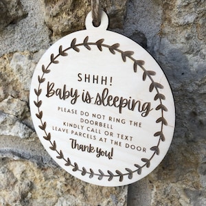 May include: A round wooden sign with a wreath design and the text "SHHH! Baby is sleeping. Please do not ring the doorbell. Kindly call or text. Leave parcels at the door. Thank you!"