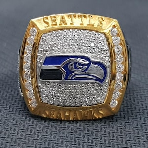 Op de afbeelding: Een goudkleurige ring met het woord "SEATTLE" bovenaan en "SEAHAWKS" onderaan. De ring heeft een blauw, zilver en zwart vogel-logo. Hij is versierd met kleine, heldere stenen. Een sieraad voor fans.