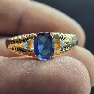 Może przedstawiać: Złoty pierścionek z dużym, owalnym, ciemnoniebieskim kamieniem szlachetnym pośrodku. Pierścionek ma wygrawerowane słowa "Riverview", "Juliana", "High" oraz rok "2025". Małe, przezroczyste kamienie osadzone są po obu stronach centralnego kamienia.