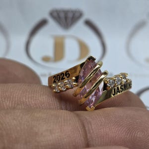 Może przedstawiać: Złoty pierścionek z różowym kamieniem centralnym w kształcie markizy i mniejszymi, przezroczystymi kamieniami. Pierścionek ma wygrawerowany napis "2026" i "Jasmine". Pierścionek jest trzymany w dłoni.