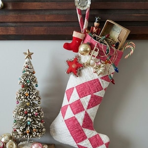 May include: A quilted Christmas stocking with a red and white geometric pattern, filled with a miniature nutcracker, a small red felt boot, a gold trumpet ornament, a star ornament, a candy cane, and a string of multicolored Christmas lights.