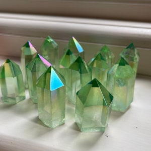 Può includere: Una collezione di punte di cristallo di quarzo aura verde. I cristalli hanno una forma geometrica sfaccettata con sfumature iridescenti sulle punte. I cristalli sono disposti su una superficie bianca.