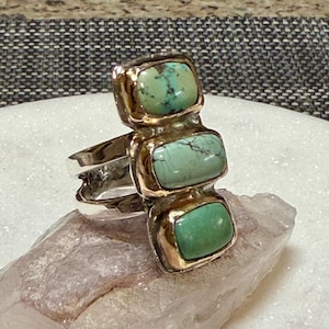 May include: A silver ring with three rectangular turquoise stones set in gold-colored bezels. The ring is displayed on a light purple crystal, showcasing the jewelry's design and color contrast.