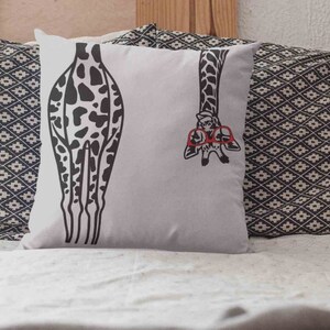 Pode incluir: Uma almofada branca com uma estampa de girafa em preto e branco. A girafa est&aacute; usando &oacute;culos vermelhos e tem a cabe&ccedil;a saindo do lado da almofada. A almofada est&aacute; em uma cama com uma colcha estampada em preto e branco.