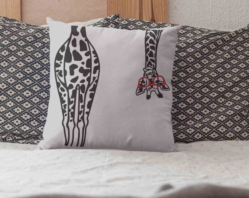 Pode incluir: Uma almofada branca com uma estampa de girafa em preto e branco. A girafa est&aacute; usando &oacute;culos vermelhos e tem a cabe&ccedil;a saindo do lado da almofada. A almofada est&aacute; em uma cama com uma colcha estampada em preto e branco.