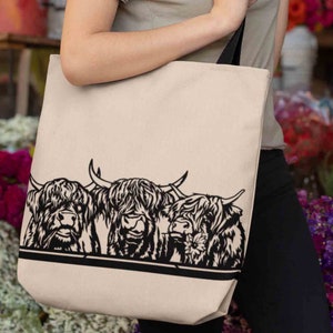 May include: A beige tote bag with black straps features a black and white illustration of three Highland cows with fluffy hair. The cows are looking at the viewer.