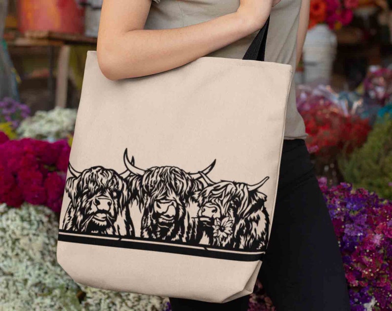 May include: A beige tote bag with black straps features a black and white illustration of three Highland cows with fluffy hair. The cows are looking at the viewer.