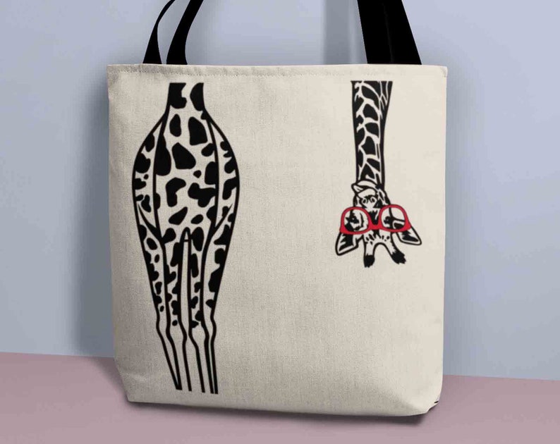 Pode incluir: Uma bolsa tote de lona branca com um design de girafa em preto e branco. A girafa est&aacute; usando &oacute;culos vermelhos e tem a cabe&ccedil;a de cabe&ccedil;a para baixo. As pernas da girafa s&atilde;o vis&iacute;veis no lado esquerdo da bolsa.