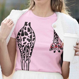 Pode incluir: Uma camiseta rosa com uma estampa de girafa em preto e branco. A girafa est&aacute; usando &oacute;culos vermelhos e tem uma girafa pequena nas costas.