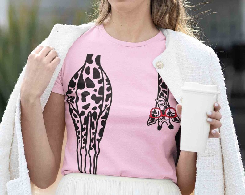 Pode incluir: Uma camiseta rosa com uma estampa de girafa em preto e branco. A girafa est&aacute; usando &oacute;culos vermelhos e tem uma girafa pequena nas costas.