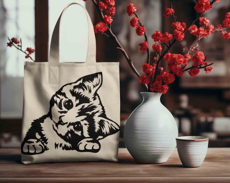 May include: A white canvas tote bag with black handles and a black and white illustration of a cat peeking out from the bottom of the bag.