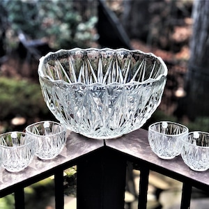 May include: A clear glass punch bowl set with a large bowl and six small square-shaped glasses. The bowl and glasses have a diamond-cut pattern. The set is displayed on a wooden surface with a blurred outdoor background.