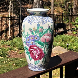 May include: A white ceramic vase with a floral design. The vase features pink, red, blue, and green flowers with green leaves. The vase has a blue and white border around the top and bottom.