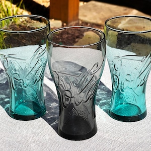 May include: Three clear Coca-Cola glasses with a teal tint. The glasses have the Coca-Cola logo embossed on the front. The glasses are on a white surface, with sunlight casting shadows.