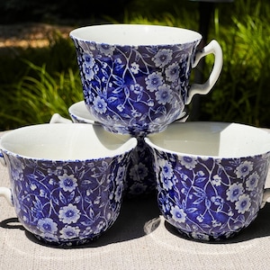 May include: Four blue and white floral teacups are stacked. The teacups have a white interior and a curved handle. The floral pattern is a detailed design of white flowers and leaves on a dark blue background. The teacups are on a light beige surface.