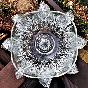 May include: Clear glass serving dish with a starburst pattern in the center and a square shape with rounded corners. The dish has decorative, petal-shaped extensions around the rim. The glass is transparent, allowing light to pass through.