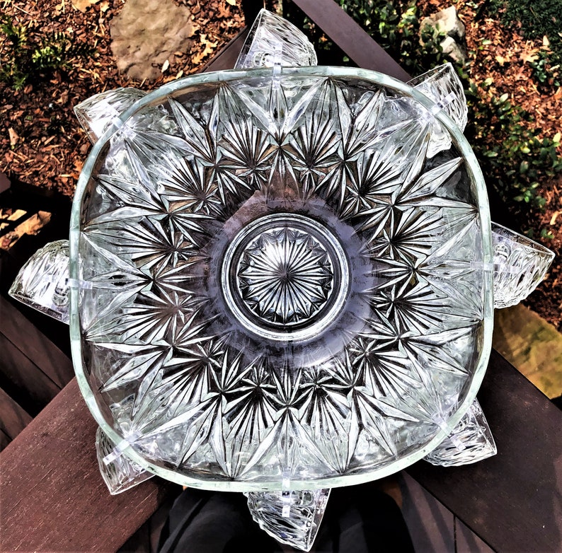 May include: Clear glass serving dish with a starburst pattern in the center and a square shape with rounded corners. The dish has decorative, petal-shaped extensions around the rim. The glass is transparent, allowing light to pass through.