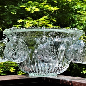 May include: A clear glass punch bowl with a scalloped rim and a grapevine design. Six matching glass cups hang from the bowl.