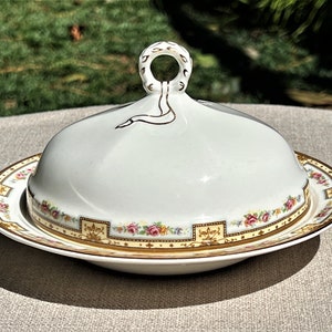 May include: A white porcelain butter dish with a lid and a floral design. The lid has a gold handle and a gold ribbon design. The dish has a gold rim and a floral design around the edge.
