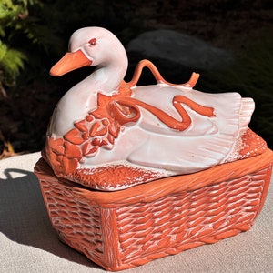 May include: A white ceramic duck with a red ribbon around its neck sits on top of a brown ceramic basket. The duck and basket are both decorated with a floral design.