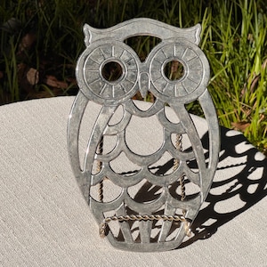 May include: A silver-colored metal owl-shaped book holder. The owl has large, round eyes and a decorative pattern on its body. A gold-colored chain supports the book holder. The item is sitting on a light-colored surface.