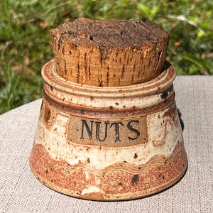 May include: A ceramic jar with a cork lid, labelled "NUTS". The jar has a beige base with reddish-brown accents and a textured cork stopper. The text "NUTS" is in black letters on a rectangular label.